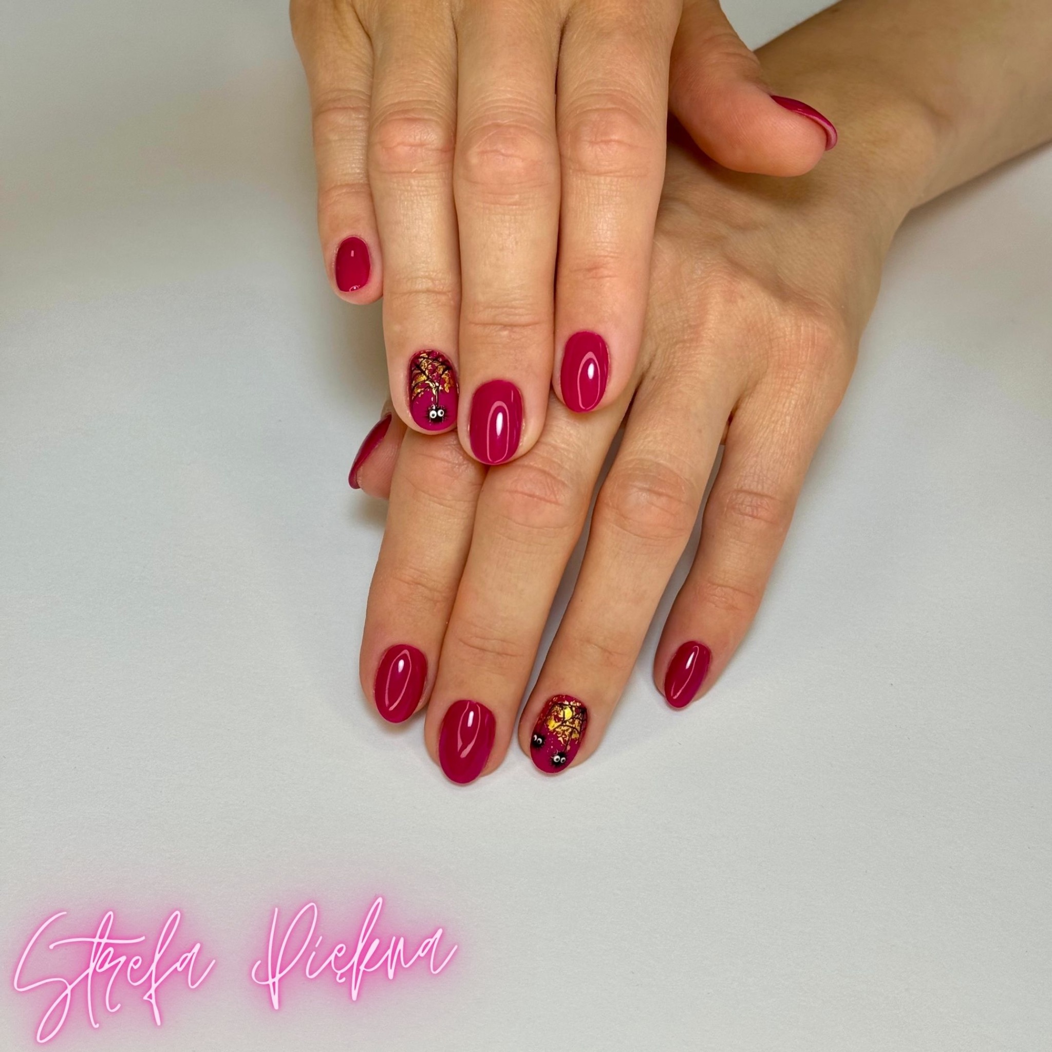 Dłonie z bordowymi paznokciami, dwa z nich ozdobione złotymi płatkami i czarnymi oczkami, na białym tle z neonowym napisem 'Strefa Piękna'.