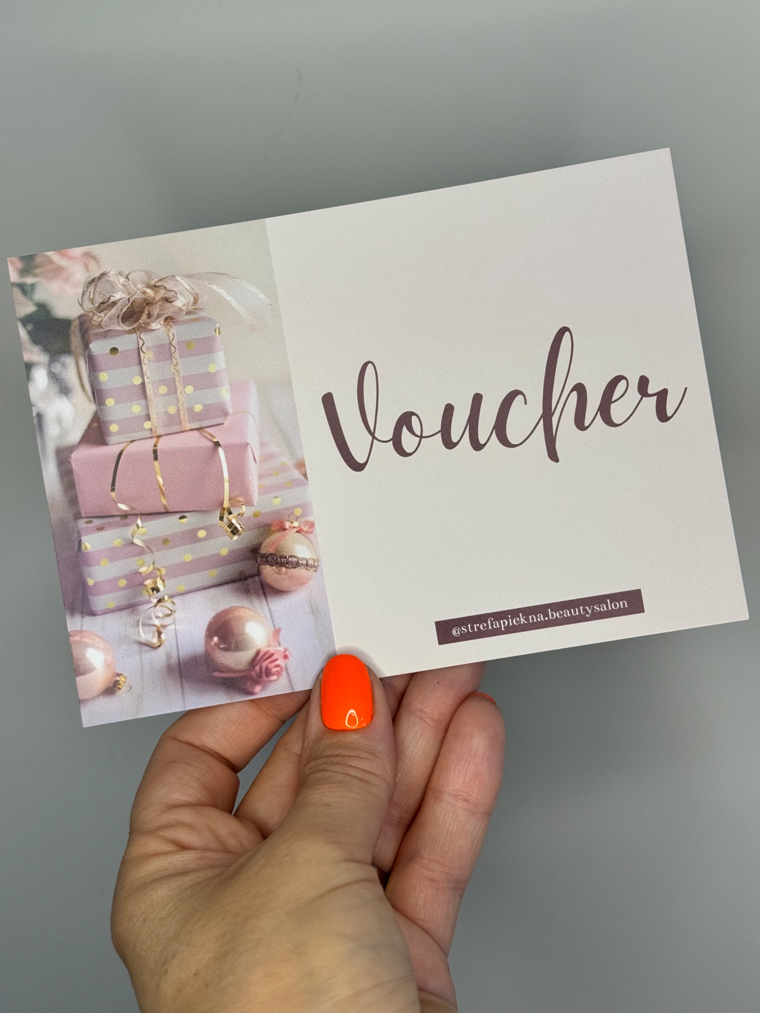 Dłoń trzymająca voucher do salonu kosmetycznego Strefa Piękna z motywem prezentów i napisem Voucher, na palcu pomarańczowy lakier hybrydowy z efektem lustra.