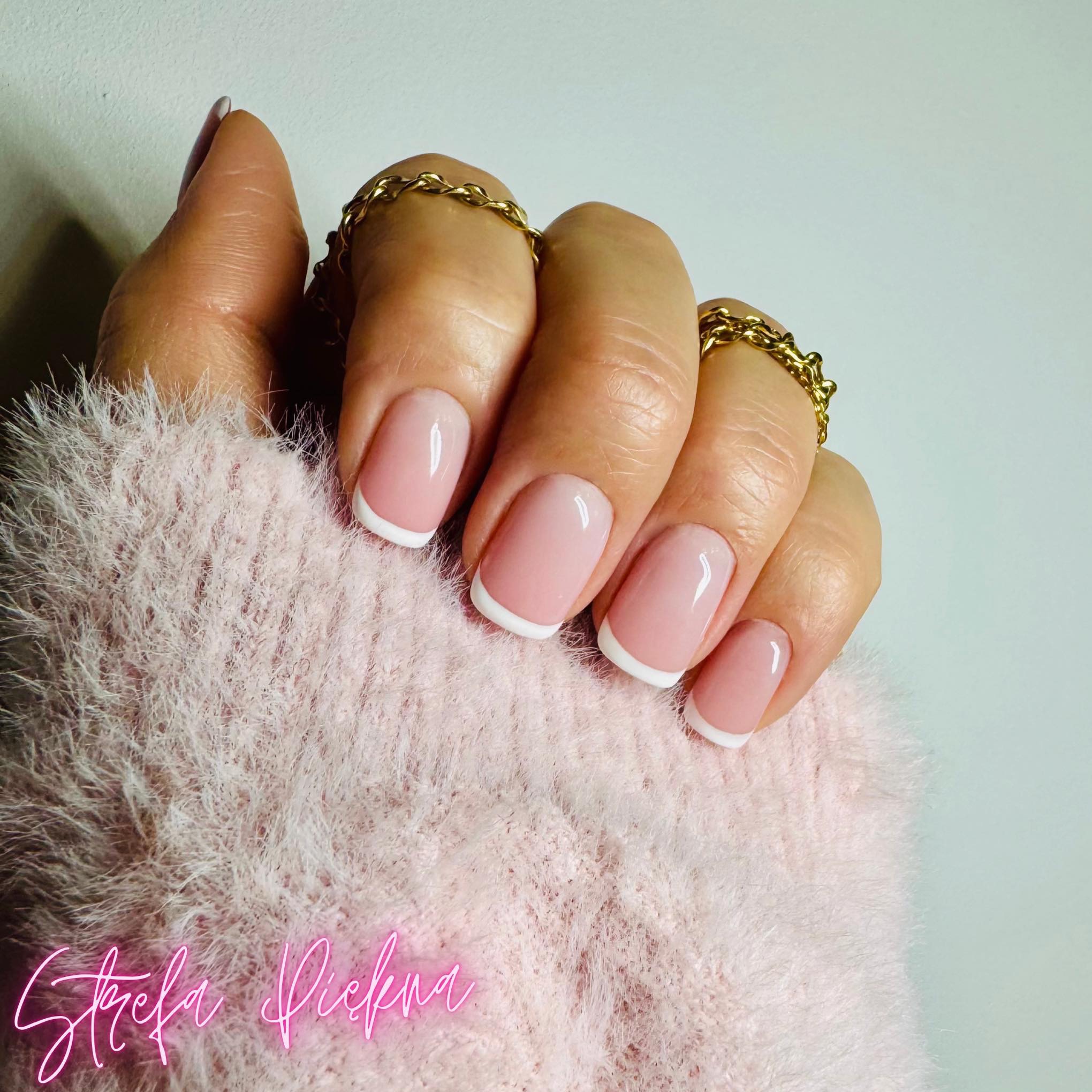 Delikatny manicure francuski na dłoni z pierścionkami w Bydgoszczy, prezentowany na tle puszystego, różowego swetra. Subtelne zdobienie paznokci.