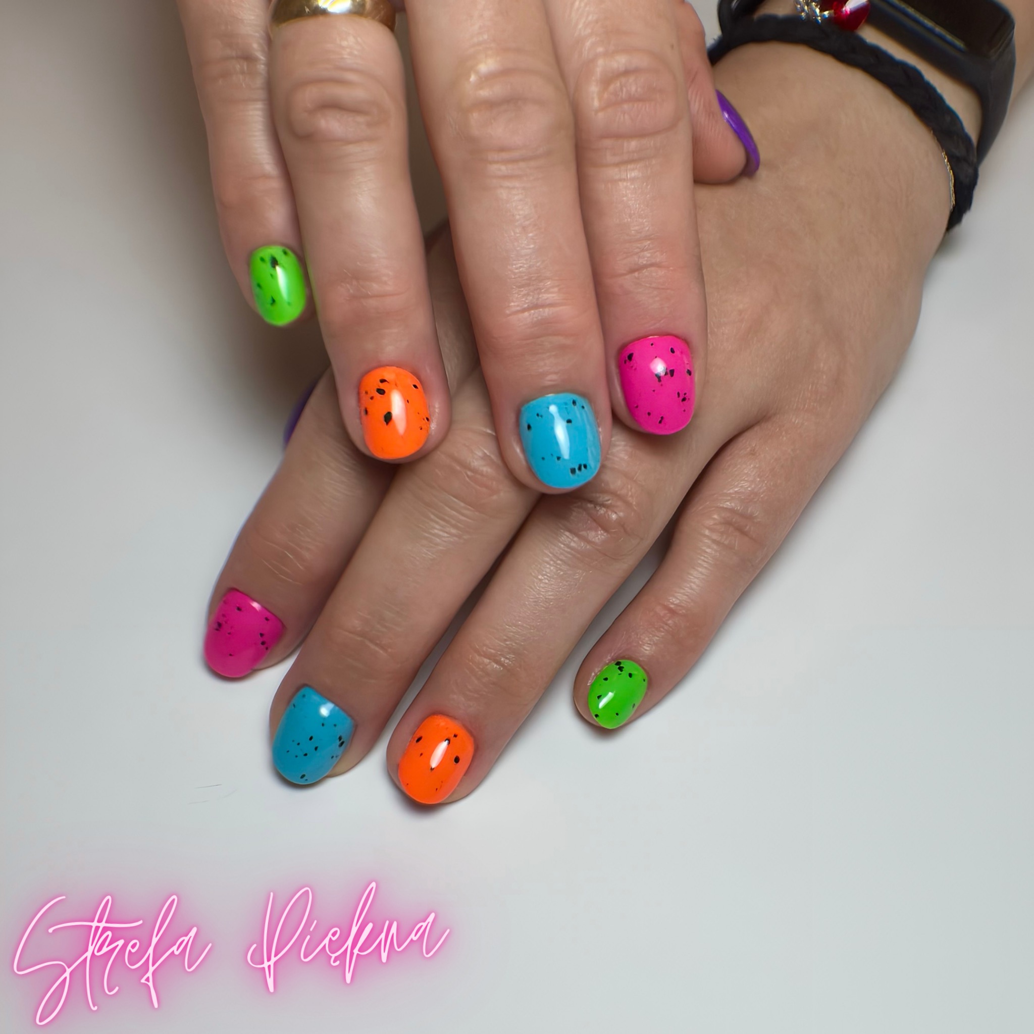 Dłonie z pomalowanymi na neonowe kolory paznokciami w kropki, na białym tle. Widoczny zegarek na ręce i sygnet na palcu. Znak wodny salonu.