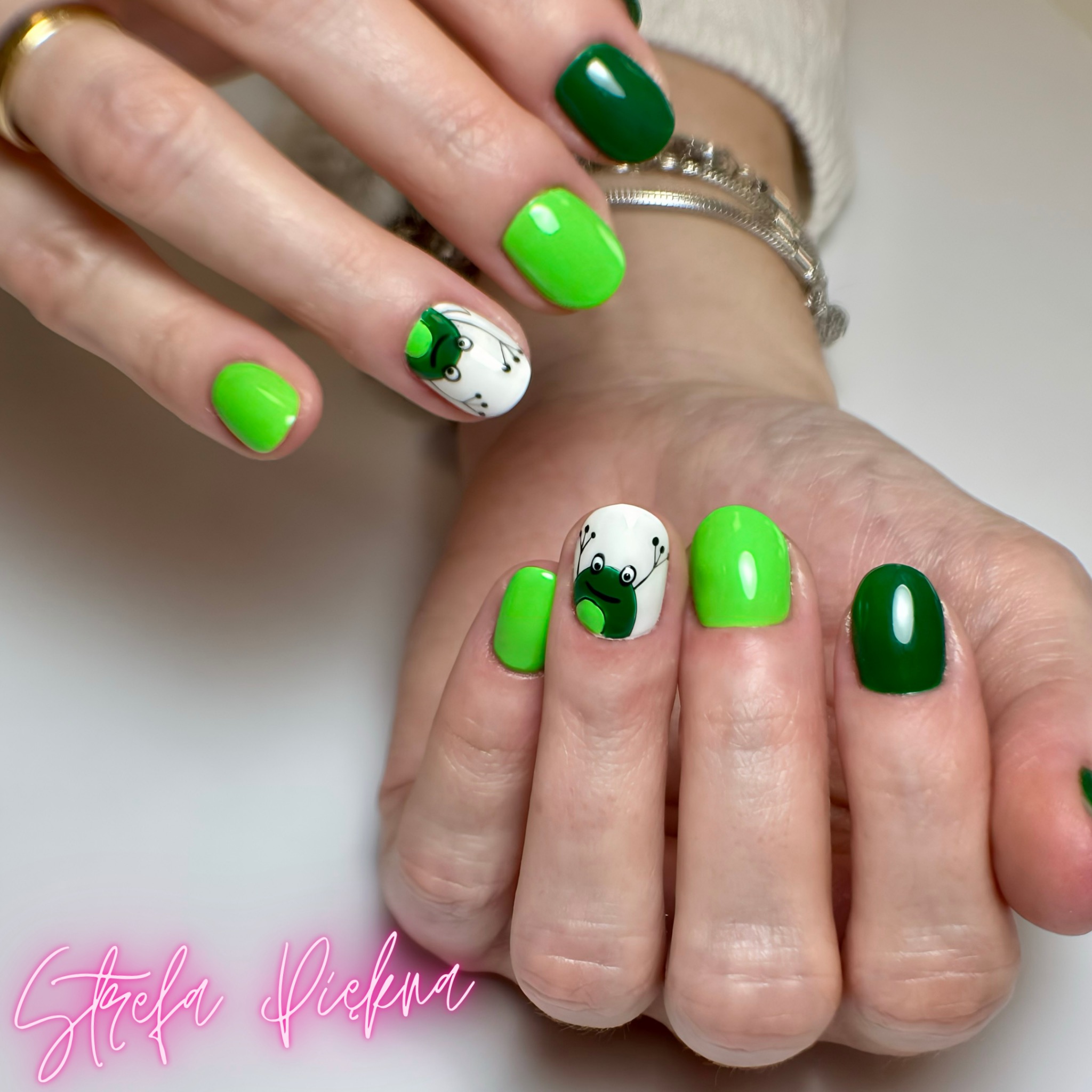 Żywe, zielone paznokcie z motywem żabki, wykonane w salonie kosmetycznym. Detal: srebrna bransoletka na nadgarstku.