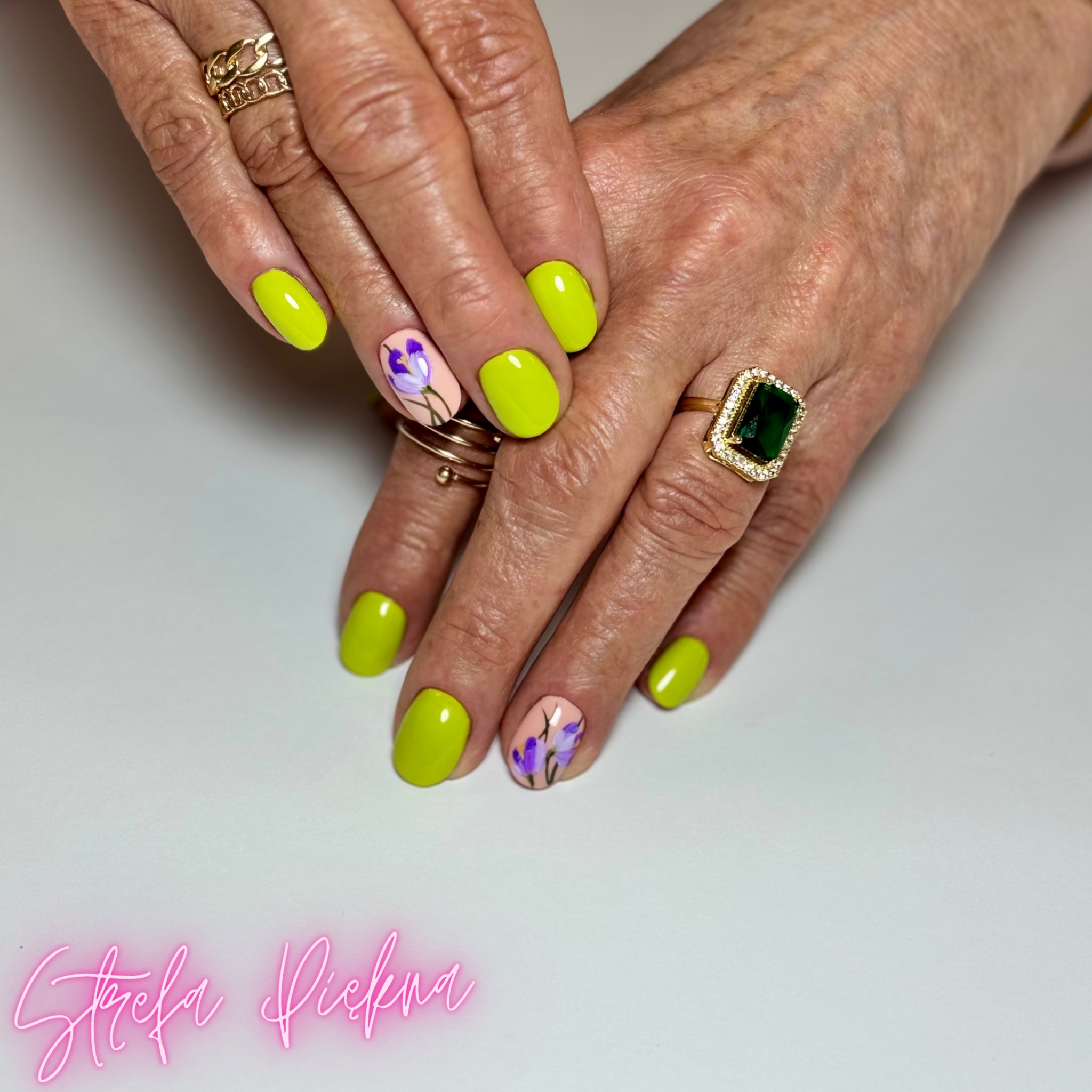 Dłonie z neonowymi, żółtymi paznokciami i kwiatowym zdobieniem na jasnym tle. Widoczne pierścionki na palcach.
