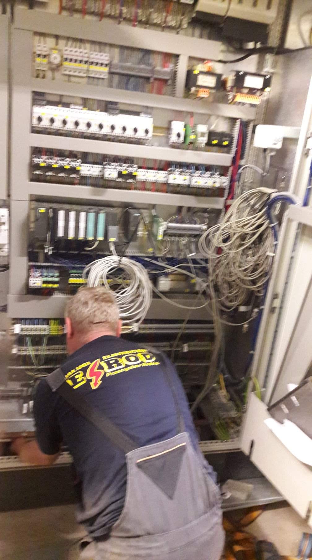 Pracownik w kombinezonie z logo firmy pracuje nad rozbudowaną szafą sterowniczą z licznymi przewodami i zabezpieczeniami.