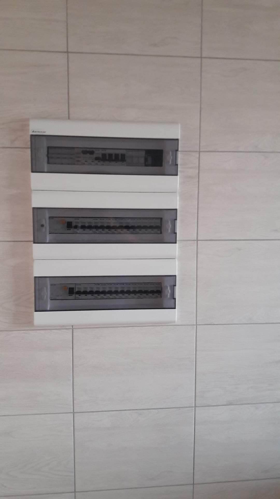 Trzy białe skrzynki rozdzielcze zamontowane na ścianie wyłożonej jasnymi, kwadratowymi płytkami imitującymi drewno.