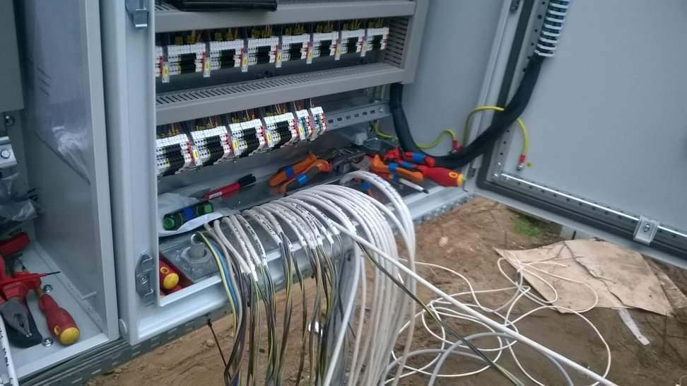 Otwarta szafa sterownicza z widocznymi listwami zaciskowymi, przewodami oznaczonymi etykietami, narzędziami i poziomnicą, umieszczona na zewnątrz budynku.