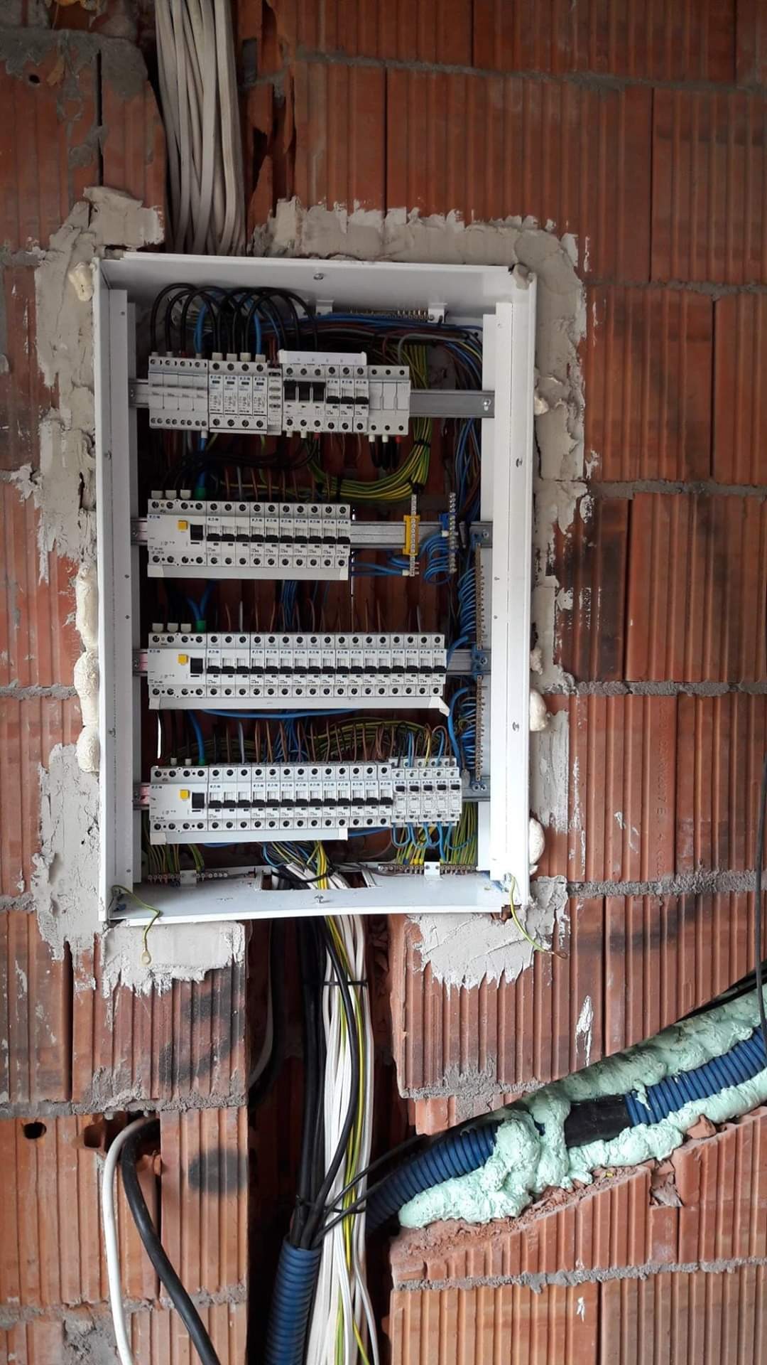 Rozdzielnica elektryczna z zabezpieczeniami w trakcie montażu w surowej ścianie z cegły, widoczne kable i pianka montażowa.