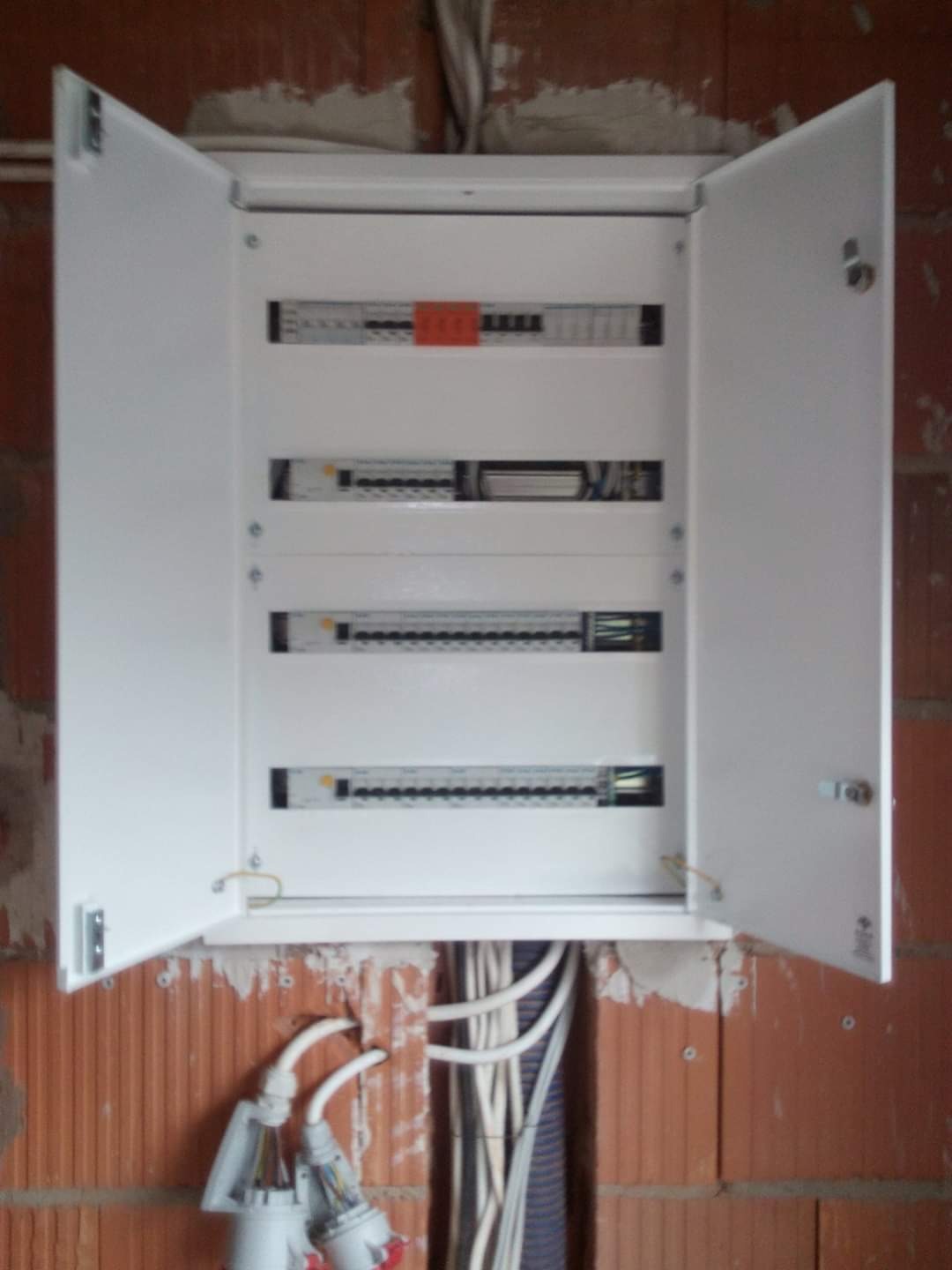Otwarta, biała skrzynka rozdzielcza z widocznymi bezpiecznikami i przewodami, zamontowana na surowej, ceglanej ścianie w trakcie instalacji.