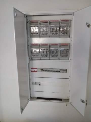 Otwarta szafka elektryczna z dwoma rzędami liczników i zabezpieczeń, widoczne białe drzwi szafki i fragment białej ściany.