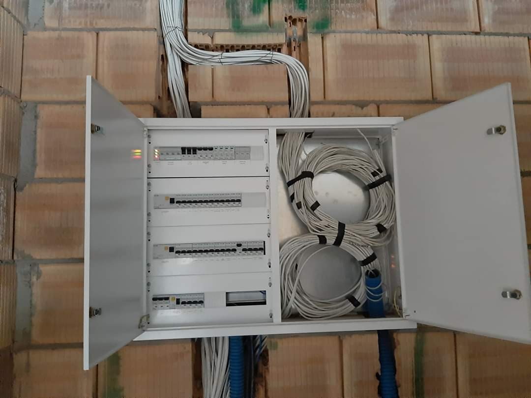 Otwarta skrzynka rozdzielcza z zamontowanymi modułami i zwiniętymi kablami w środku, widoczna instalacja w ścianie z pustaków ceramicznych i peszle z niebieskimi kablami.