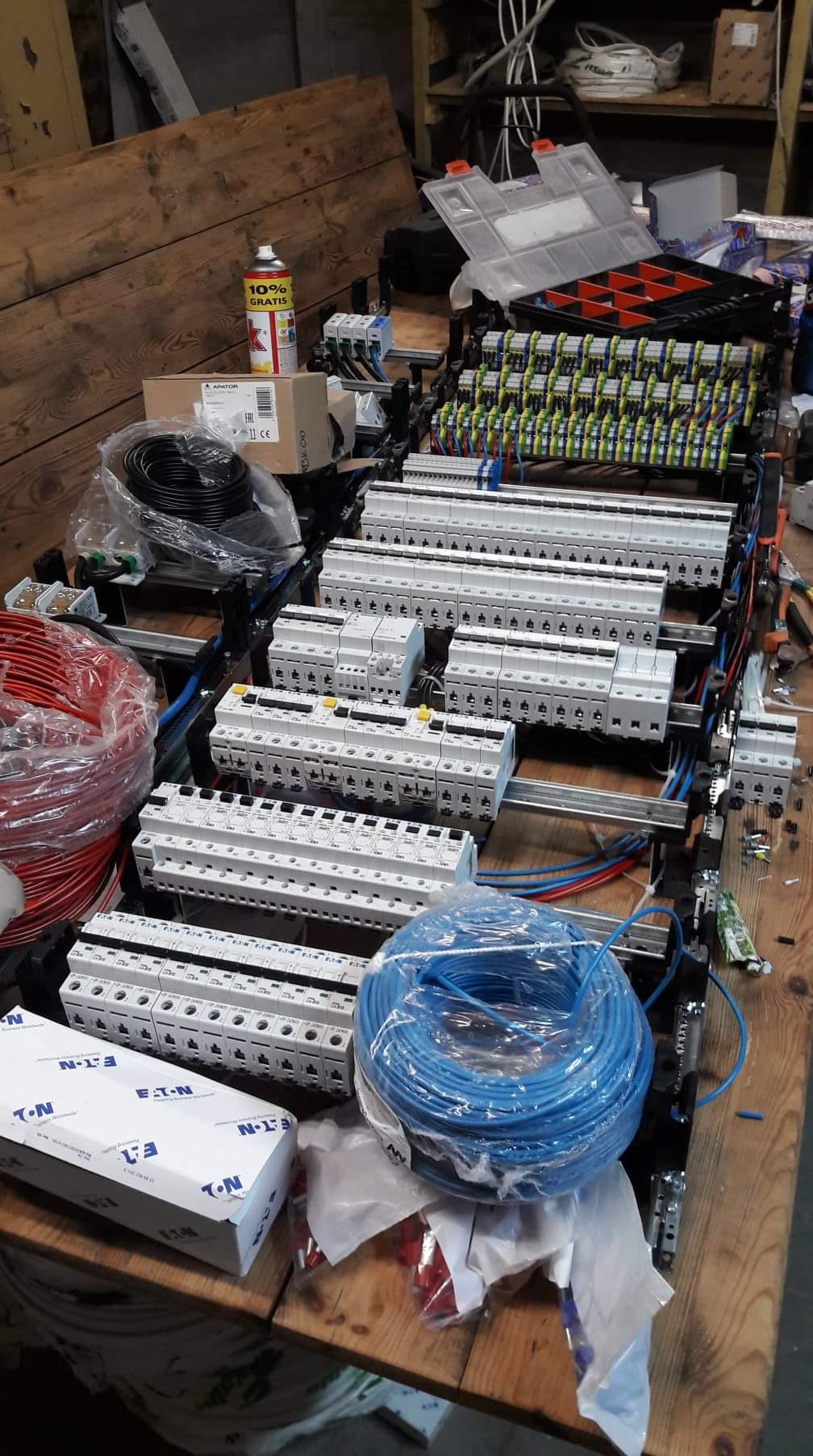 Rozdzielnica elektryczna w trakcie montażu, widoczne zabezpieczenia modułowe, przewody w różnych kolorach, puszka z narzędziami i puszka farby w tle, na drewnianym stole.