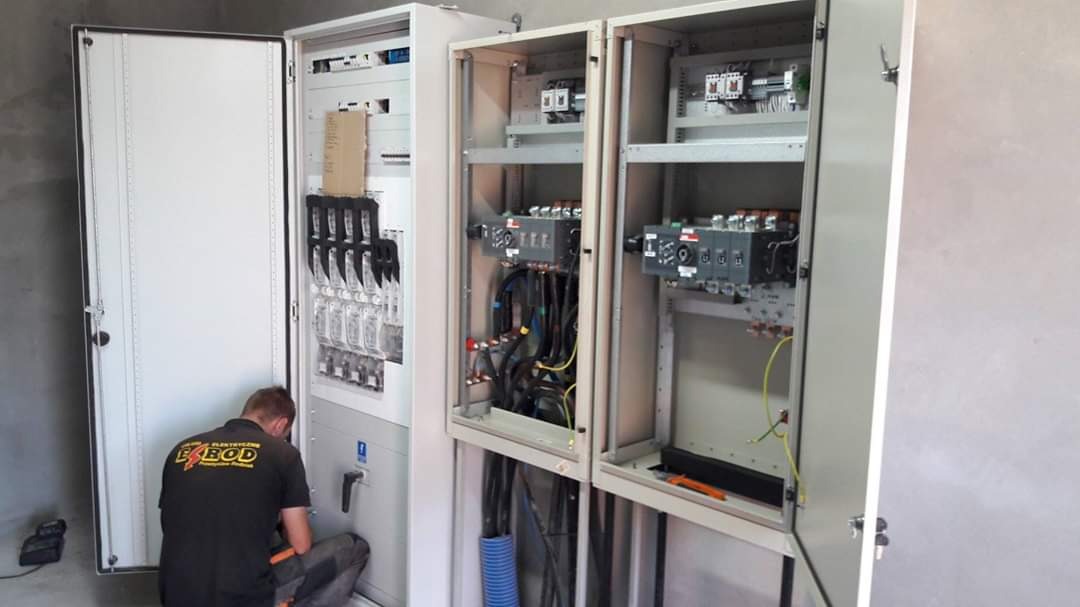Mężczyzna w czarnej koszulce z logiem firmy pracuje nad rozbudowaną rozdzielnicą elektryczną; widoczne otwarte szafy sterownicze z okablowaniem i zabezpieczeniami.