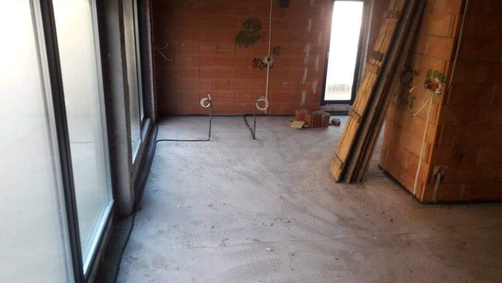 Surowy stan wnętrza w Kaliszu przed wykończeniem: widoczne przewody elektryczne wystające z podłogi, ceglana ściana z instalacją elektryczną i okna z częściowo zamontowanymi ramami.