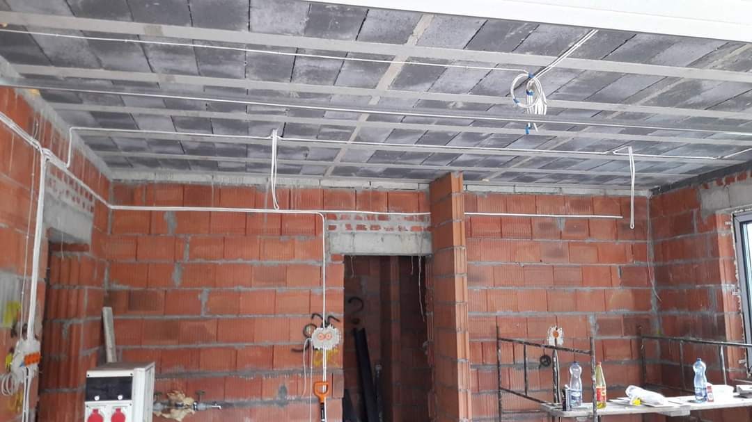 Surowy stan wnętrza z częściowo poprowadzonymi przewodami elektrycznymi po ścianach z czerwonej cegły i pod stropem z szarych płyt.