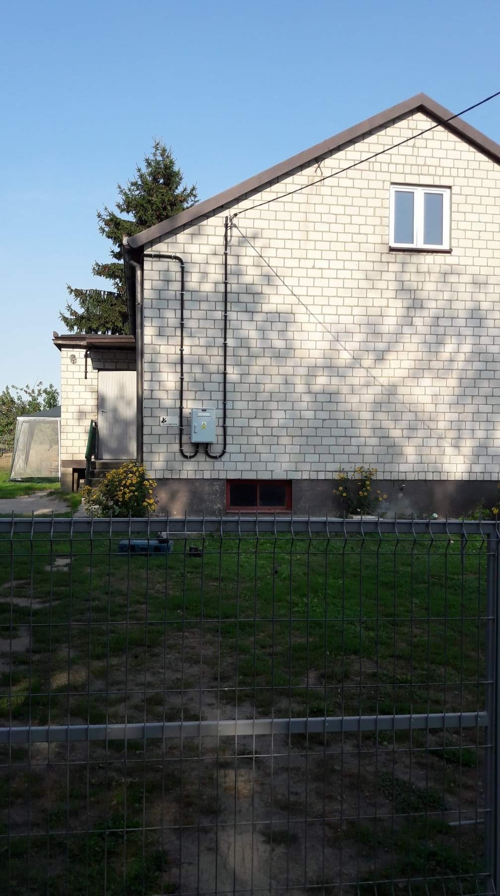 Biały murowany dom z widoczną instalacją elektryczną na zewnątrz, poprowadzoną w czarnych rurach do szarej skrzynki elektrycznej, widziany zza metalowego ogrodzenia.