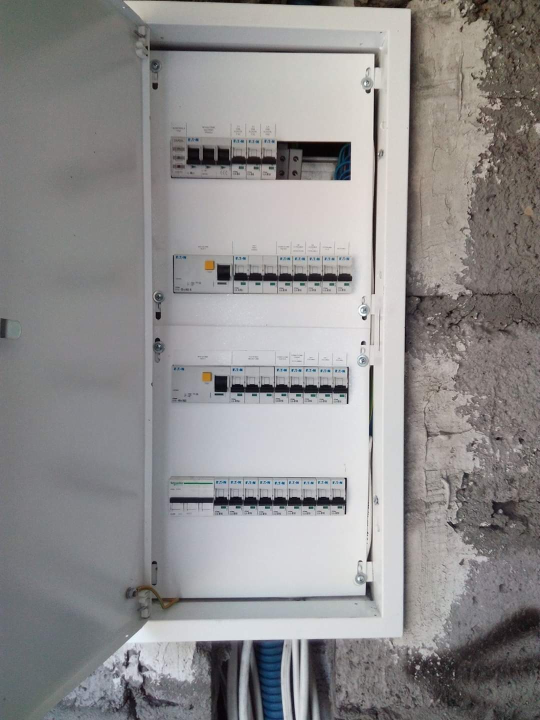 Otwarta skrzynka rozdzielcza z bezpiecznikami Eaton i Schneider na tle surowej ściany z widocznymi przewodami w peszlach.