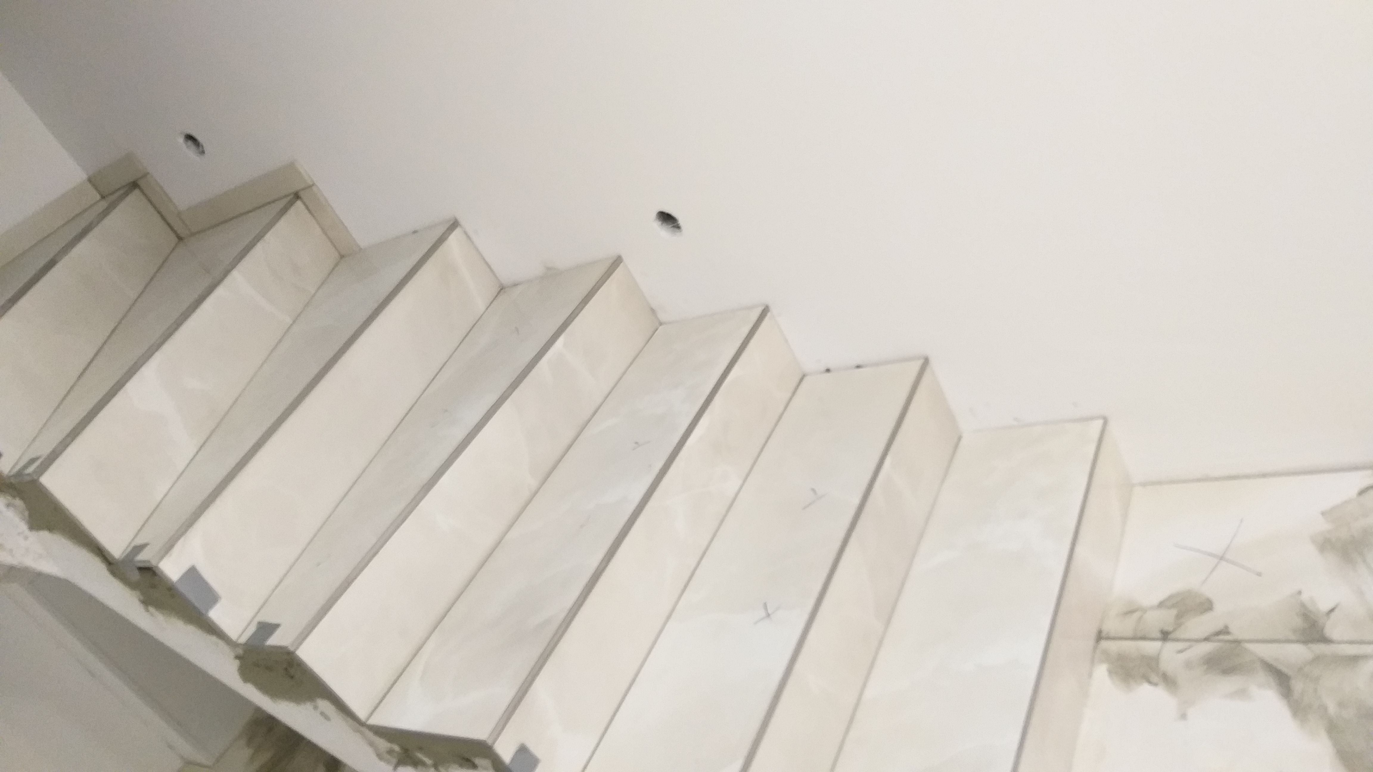 Wykończone stopnie schodów wyłożone jasnymi płytkami imitującymi marmur, widoczne ślady zaprawy i oznaczenia montażowe na ścianie.
