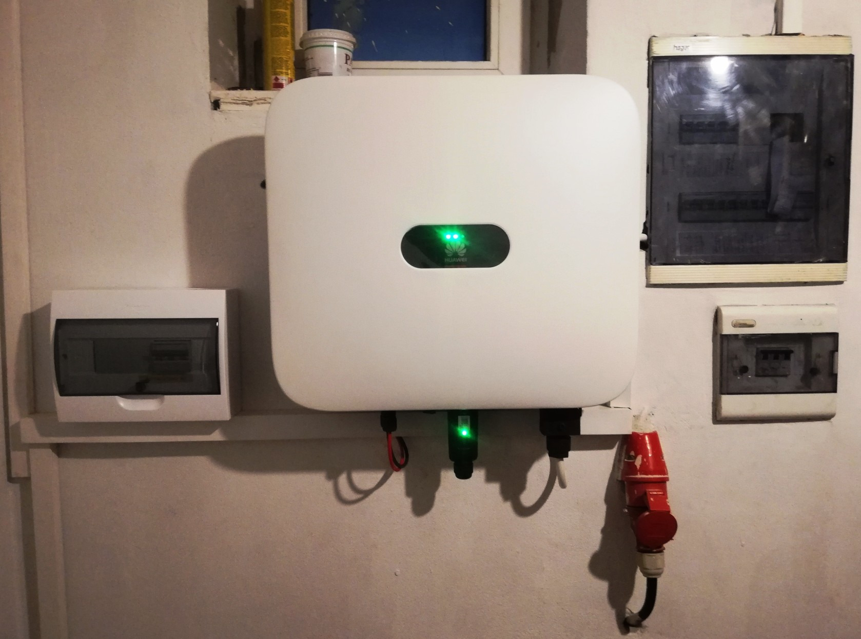 Biały inwerter fotowoltaiczny Huawei z zielonymi diodami LED, zamontowany na ścianie obok skrzynek elektrycznych, pod oknem z widocznymi puszkami farb.