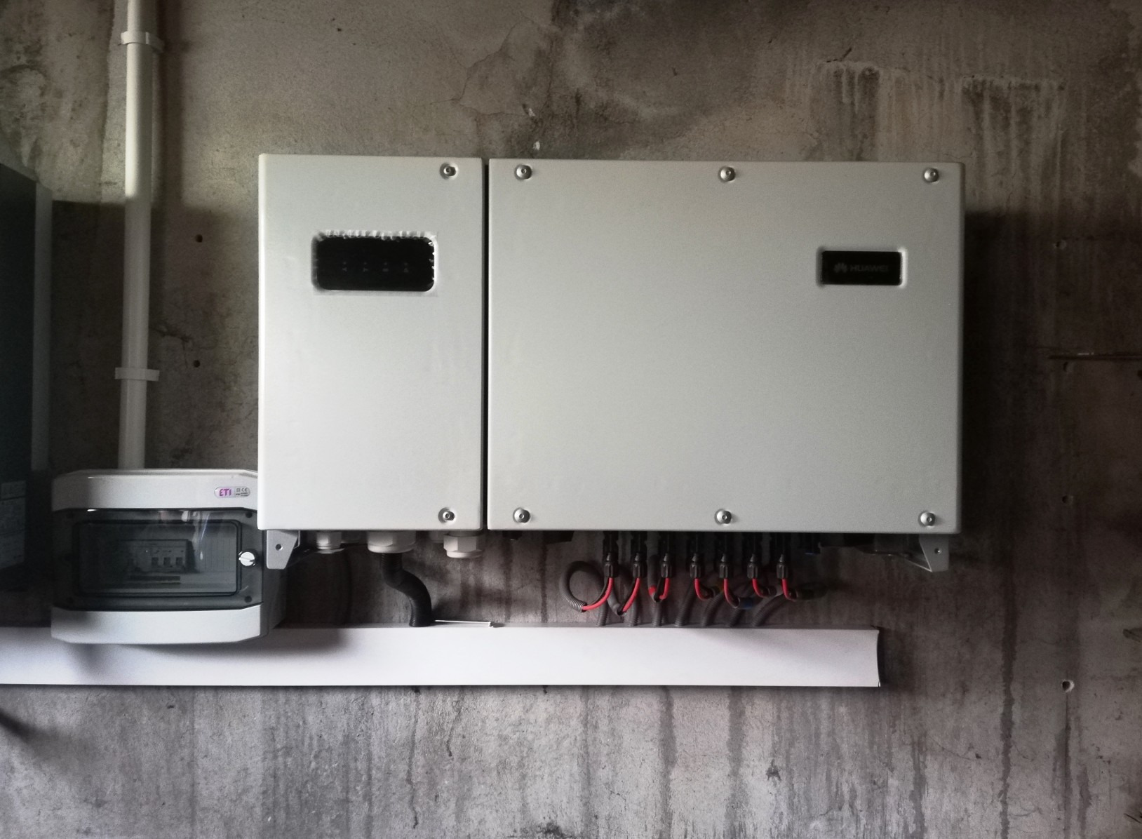 Szary falownik fotowoltaiczny marki Huawei zamontowany na surowej, betonowej ścianie, widoczne przewody i zabezpieczenie ETI.