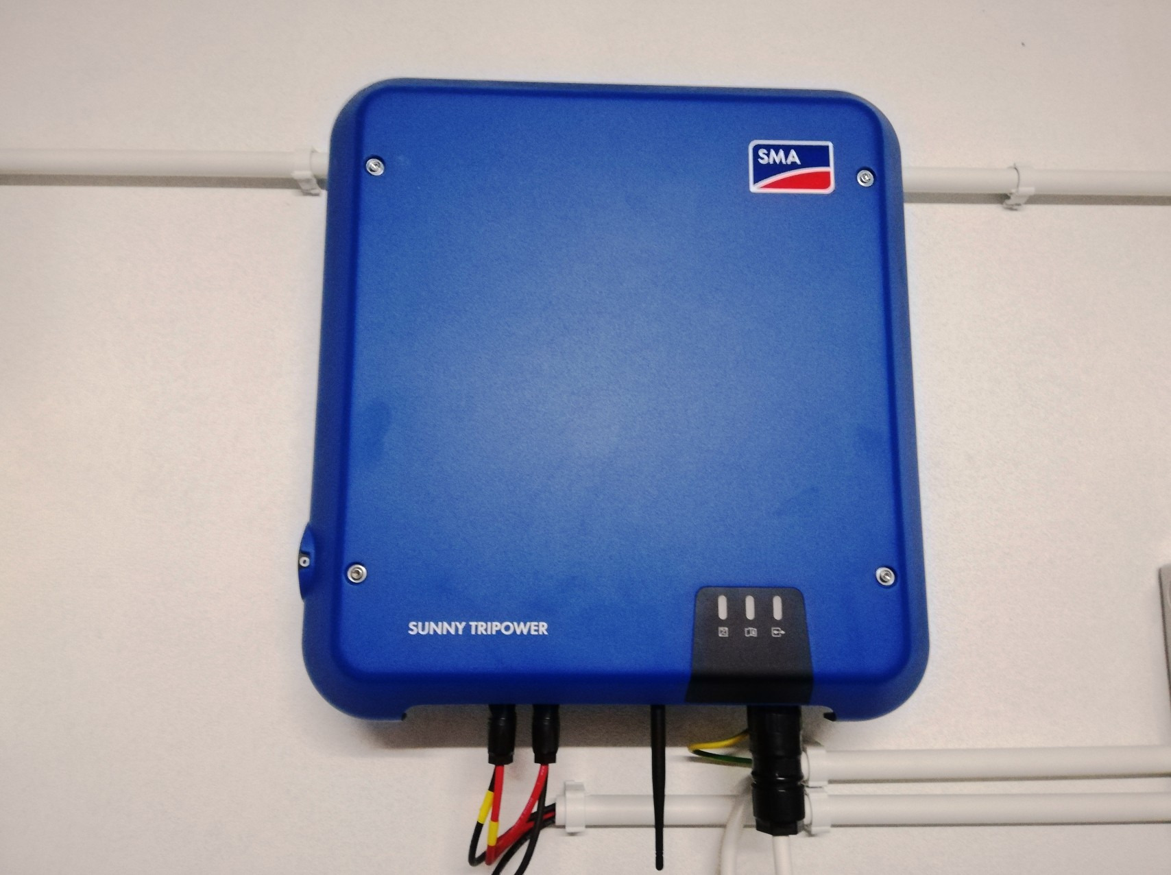 Niebieski inwerter fotowoltaiczny SMA Sunny Tripower zamontowany na białej ścianie, z widocznymi kablami i trzema diodami LED sygnalizującymi stan pracy.