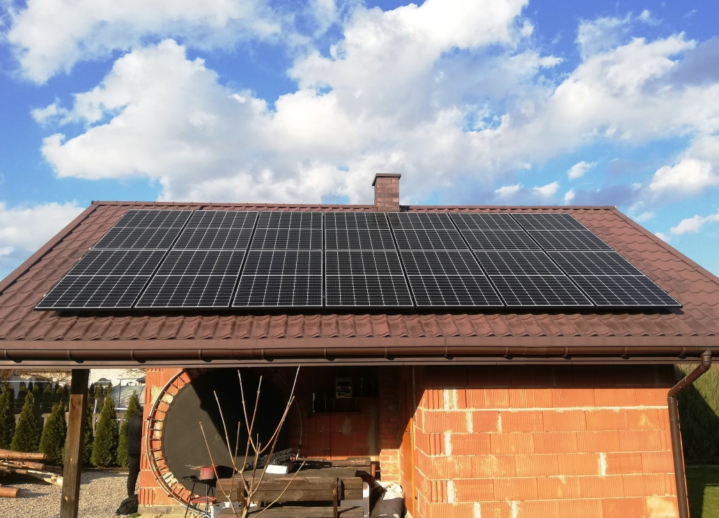 Panele fotowoltaiczne zamontowane na brązowym dachu budynku z cegły, widoczne błękitne niebo z chmurami w tle.