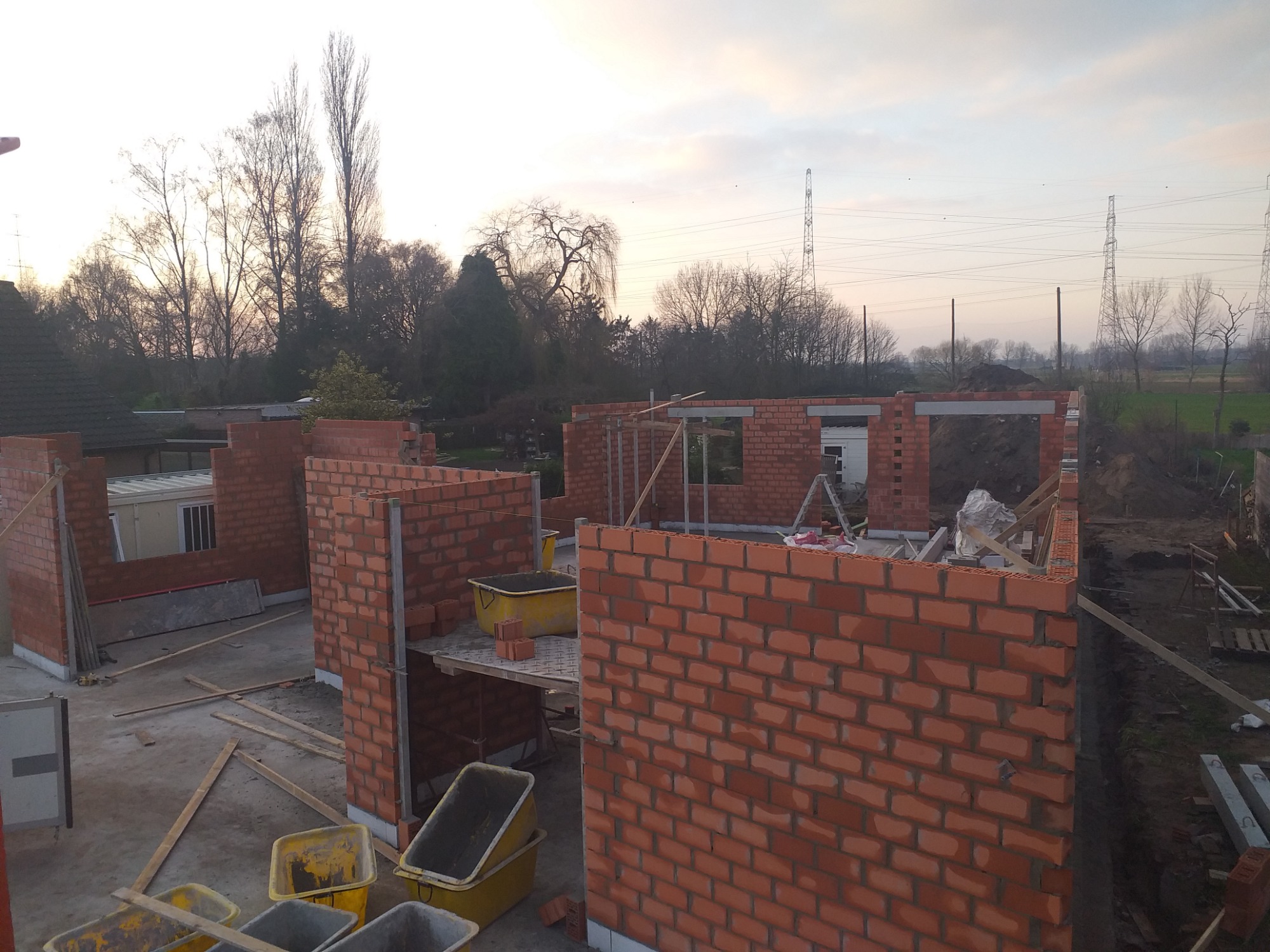 Wznoszenie ścian z czerwonej cegły na budowie domu w Kamiennej Górze, widoczne żółte pojemniki na zaprawę, w tle drzewa i słupy wysokiego napięcia.