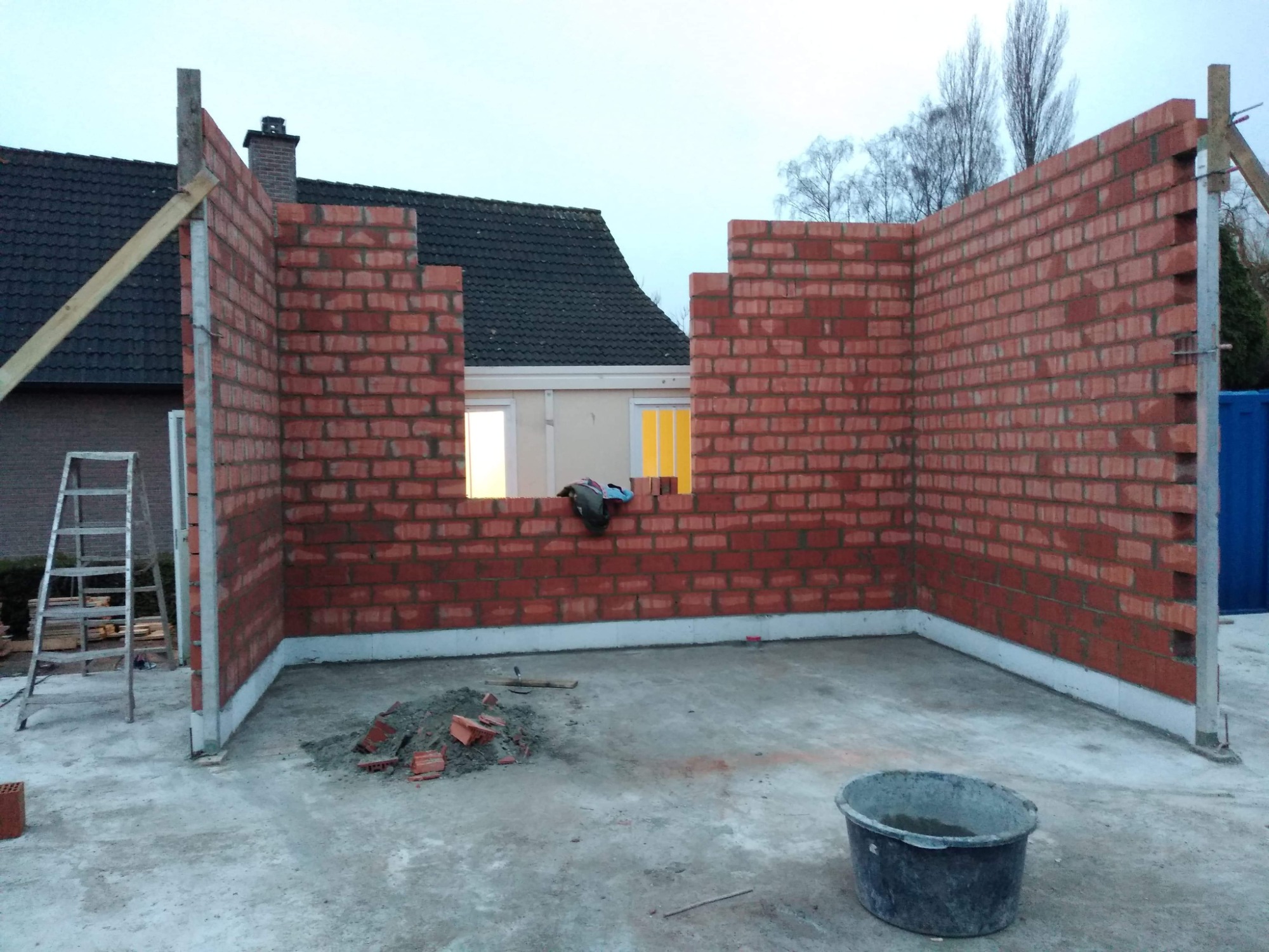 Wznoszone ściany z czerwonej cegły na fundamencie, widoczne okno w ścianie, drabina oparta o ścianę, wiadro z zaprawą na posadzce, fragment dachu domu w tle.