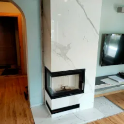 Biały kominek z czarną ramą, wykończony płytami imitującymi marmur, w trakcie montażu, widoczne niedociągnięcia wykończeniowe przy ścianie i podłodze.