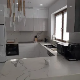 Nowoczesna kuchnia z białymi, lakierowanymi frontami szafek, blatem kamiennym z wyraźnym żyłkowaniem imitującym marmur, oraz wiszącą lampą z podłużnymi, szklanymi elementami.