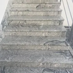 Bieg schodów z szarego kamienia z ciemniejszymi żyłkami, zabezpieczony metalową barierką po prawej stronie, widok z dołu.