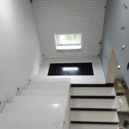 Nowoczesne białe schody z podświetleniem LED w ścianie, prowadzące do czarnego panelu dekoracyjnego pod oknem dachowym, widok z dołu.