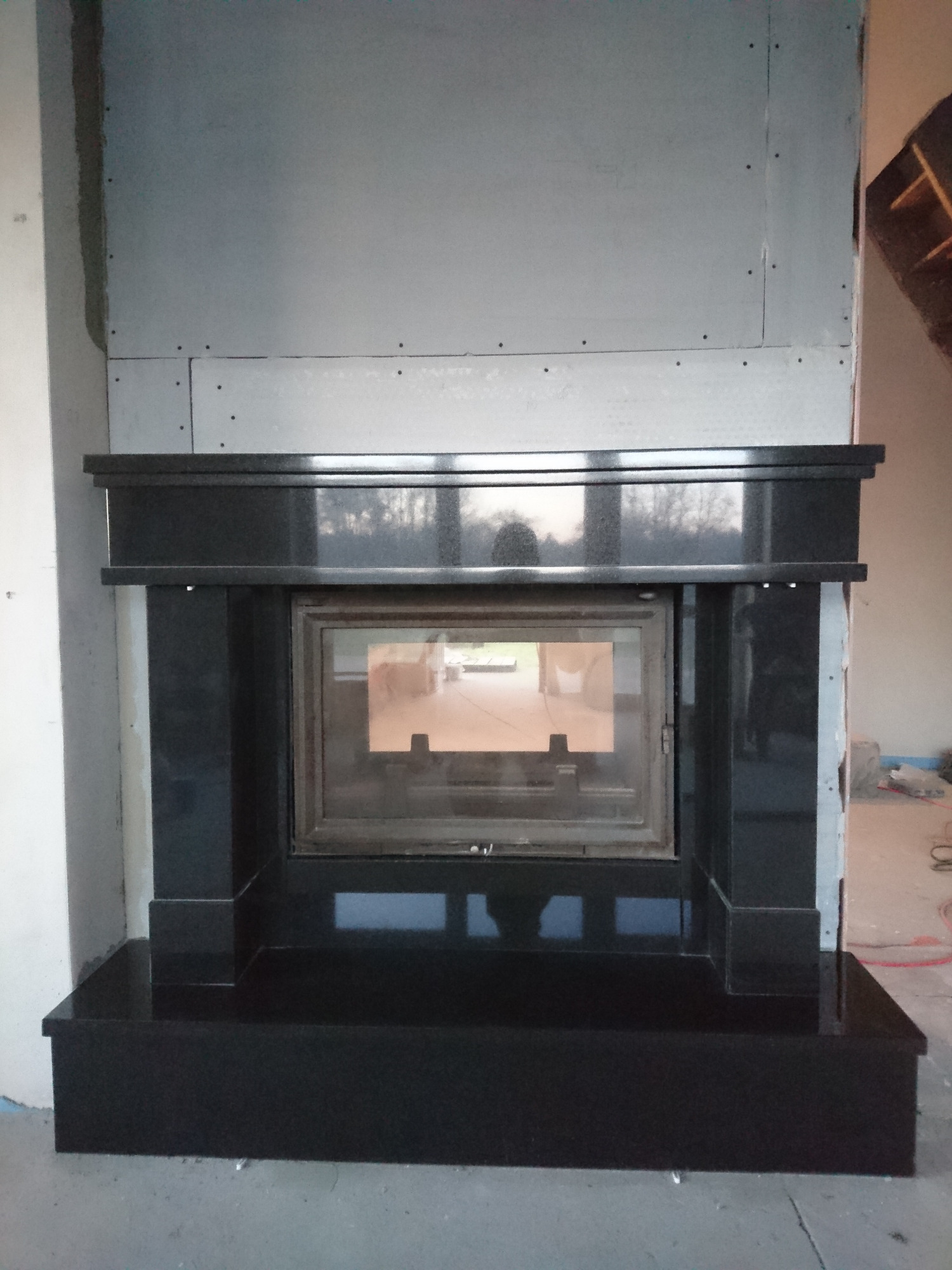 Nowoczesna obudowa kominka z czarnego kamienia, widoczna w trakcie montażu na tle surowych ścian z płyt gipsowo-kartonowych i elementami instalacji elektrycznej na podłodze.