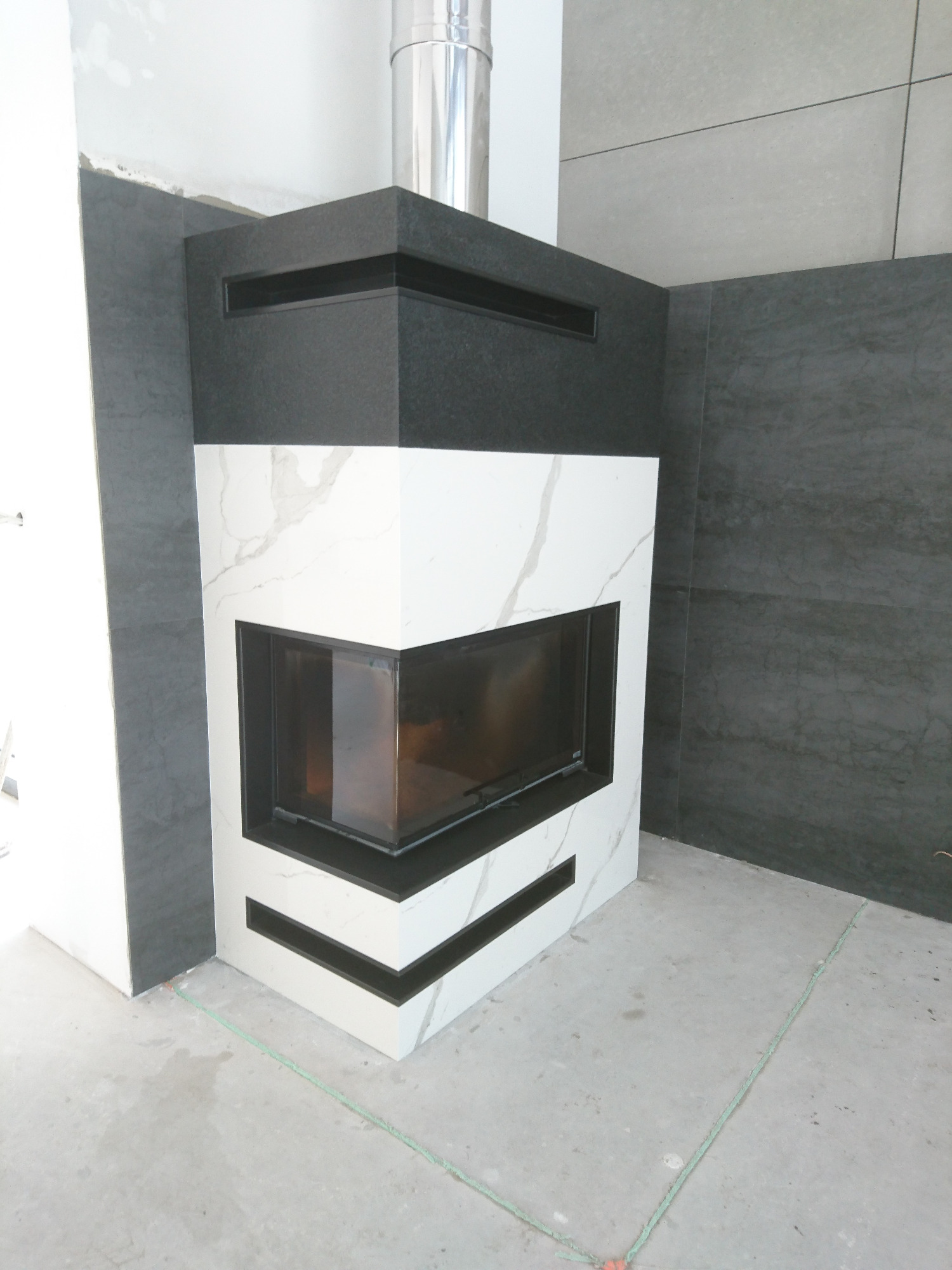 Nowoczesny kominek z obudową w kolorach czerni i bieli, z elementami imitującymi marmur, widoczny w trakcie montażu, z szarą płytką na ścianie w tle.