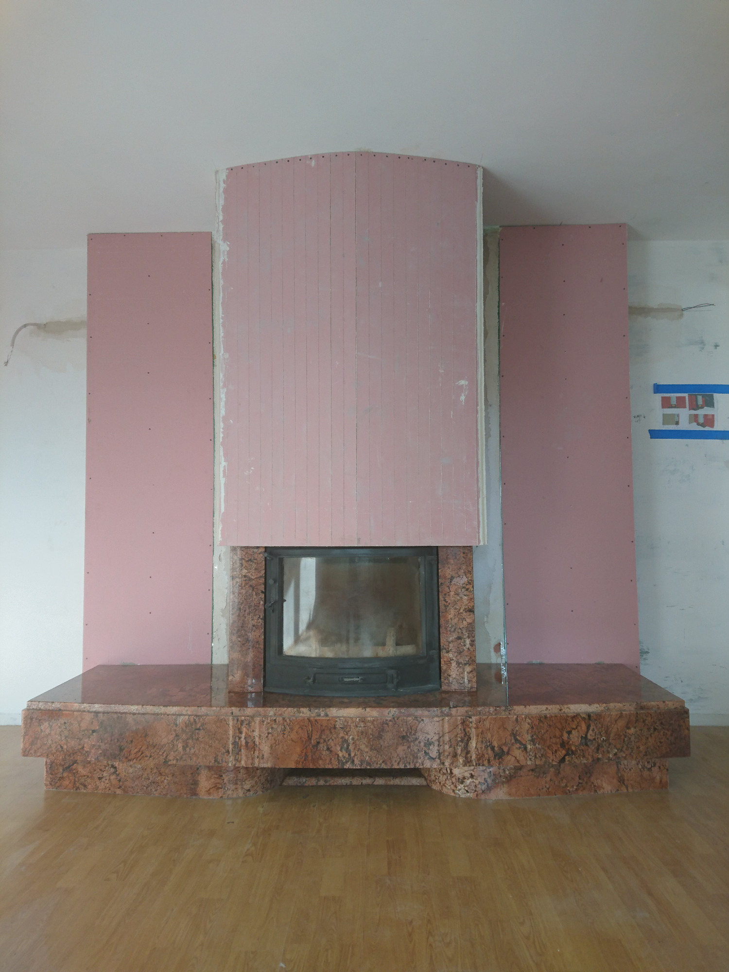 Kominek z obudową z czerwonego kamienia w trakcie montażu, widoczne płyty kartonowo-gipsowe w kolorze różowym i fragment ściany z instalacjami.