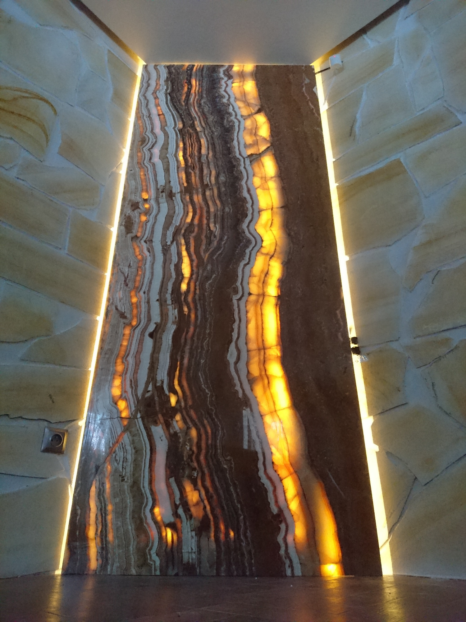 Podświetlona ściana z onyksu z naturalnymi wzorami, otoczona kamienną okładziną i oświetleniem LED po bokach, tworząca efektowny element dekoracyjny wnętrza.