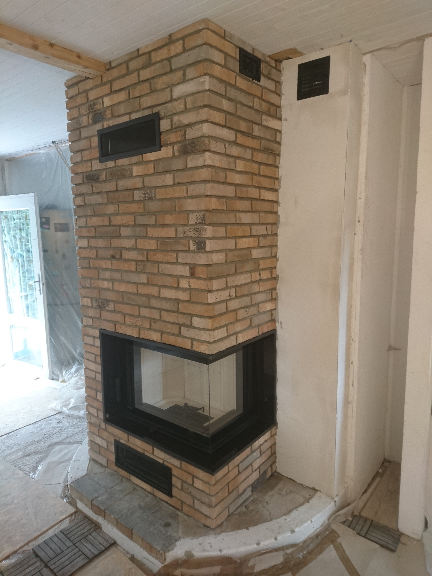 Murowany kominek z cegły klinkierowej w trakcie budowy, widoczne czarne wloty powietrza, surowy stan wykończenia wnętrza.