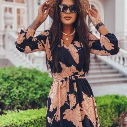 Kobieta w kwiecistej, czarno-beżowej sukience, słomkowym kapeluszu i okularach przeciwsłonecznych, stojąca na tle budynku z kolumnami i zielenią.