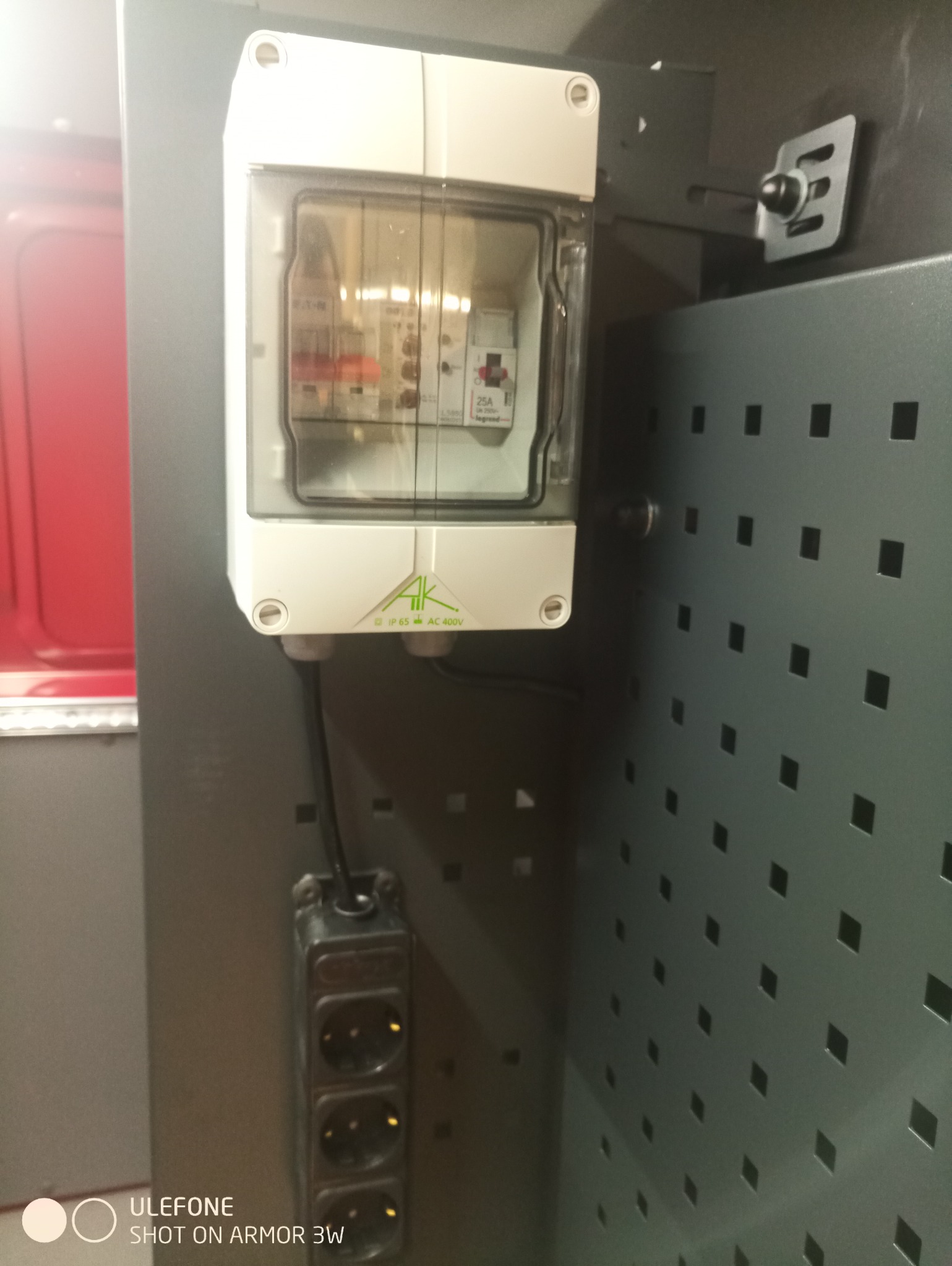 Zamknięta skrzynka elektryczna z widocznymi bezpiecznikami i listwą zasilającą, zamontowana na perforowanej ścianie wewnątrz pojazdu.