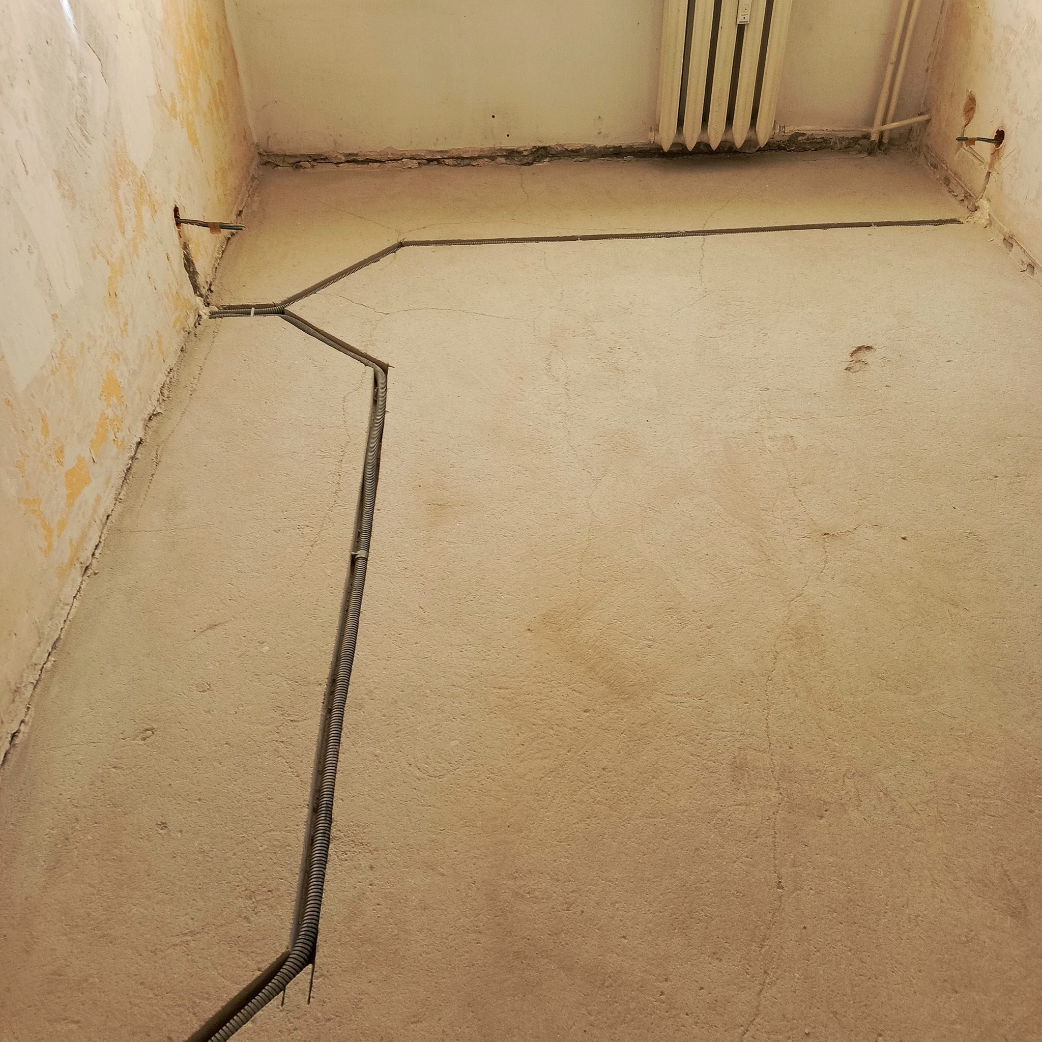 Szara, karbowana rura instalacji elektrycznej ułożona w bruzdach na ścianie, z widocznymi przewodami w otworach. W tle grzejnik.