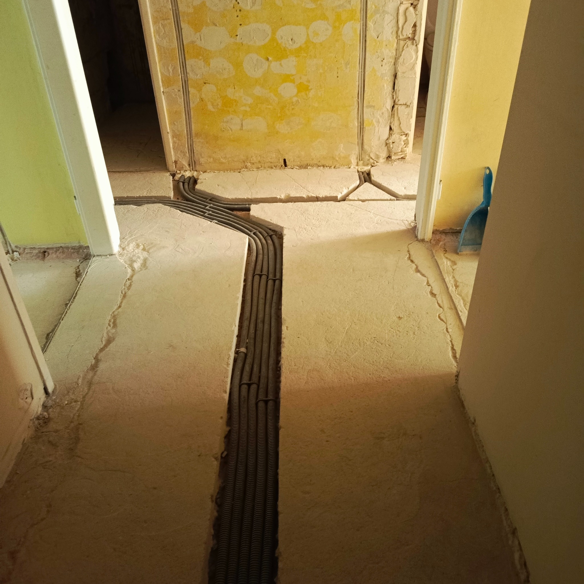 Korytarz w trakcie remontu z widocznymi rurami instalacji elektrycznej ułożonymi w bruzdach w podłodze. Ściany w stanie surowym, widoczne warstwy farb.