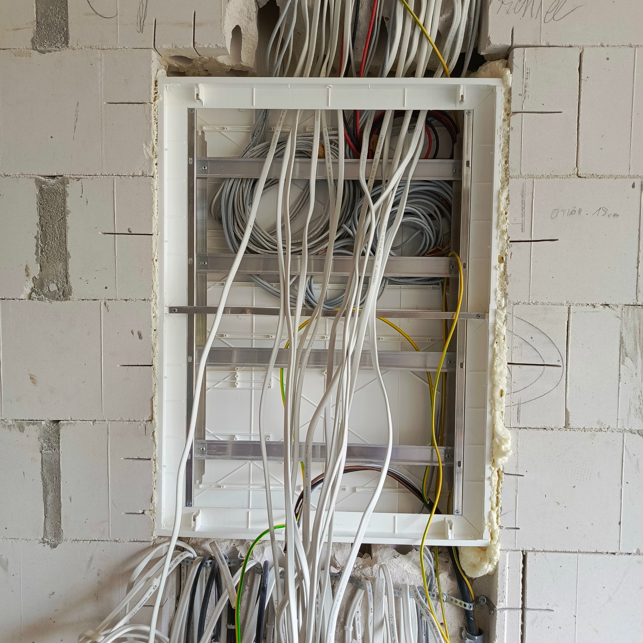 Rozdzielnica elektryczna w trakcie montażu w ścianie z betonu komórkowego. Widoczne przewody i puszka zabezpieczona pianką montażową. Etap prac instalacyjnych.