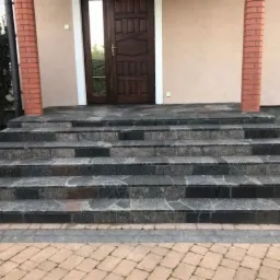 Zewnętrzne schody wejściowe do domu, wyłożone nieregularnymi płytami kamiennymi w odcieniach szarości i czerni, z widoczną kostką brukową na podejściu i ceglanymi filarami po bokach.