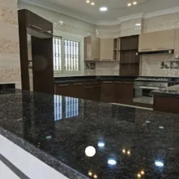 Błyszczący, ciemny blat z widocznymi drobinkami w nowocześnie urządzonej kuchni z beżowo-brązowymi szafkami i dekoracyjnymi płytkami na ścianie.