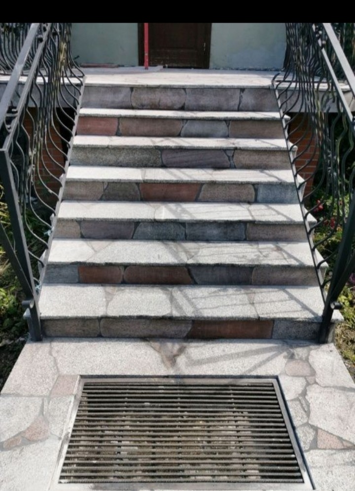 Kamienne schody wejściowe z kutą balustradą i kratką odpływową, prowadzące do ciemnych drzwi.