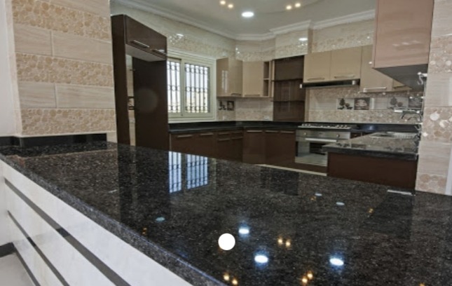 Błyszczący, ciemny blat z widocznymi drobinkami w nowocześnie urządzonej kuchni z beżowo-brązowymi szafkami i dekoracyjnymi płytkami na ścianie.