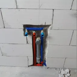Wnęka w ścianie z betonu komórkowego z fragmentami instalacji wodno-kanalizacyjnej: rury niebieskie i szare z czerwonymi złączkami, zabezpieczone pianką montażową.