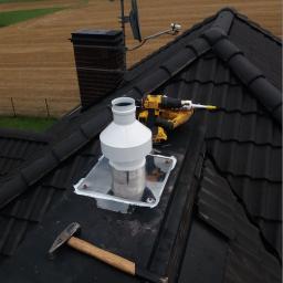 Olesek Instalacje. Projektowanie i wykonywanie instalacji sanitarnych - Montaż rekuperacji na dachu z użyciem wiertarki i młotka, widok z góry na czarną dachówkę i krajobraz polny w tle.
