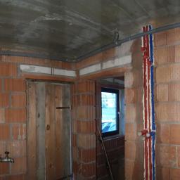 Olesek Instalacje. Projektowanie i wykonywanie instalacji sanitarnych - Wnętrze w trakcie budowy z widocznymi szarymi rurami instalacji wodnej pod sufitem, pionowymi rurami z izolacją w kolorach czerwonym i niebieskim, oraz zaworami przy podłodze na tle muru...