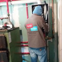 Olesek Instalacje. Projektowanie i wykonywanie instalacji sanitarnych - Instalator w niebieskiej czapce w trakcie montażu pieca CO, widoczne rury instalacji grzewczej w izolacji, oraz starszy piec w tle