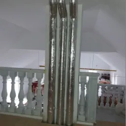 Trzy pionowe rury owinięte folią izolacyjną, umieszczone przy białej ścianie na poddaszu z balustradą w tle.