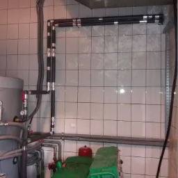 Kotłownia z widocznymi rurami instalacji grzewczej, zbiornikiem, pompą obiegową i urządzeniem spawalniczym na tle wykafelkowanej ściany.